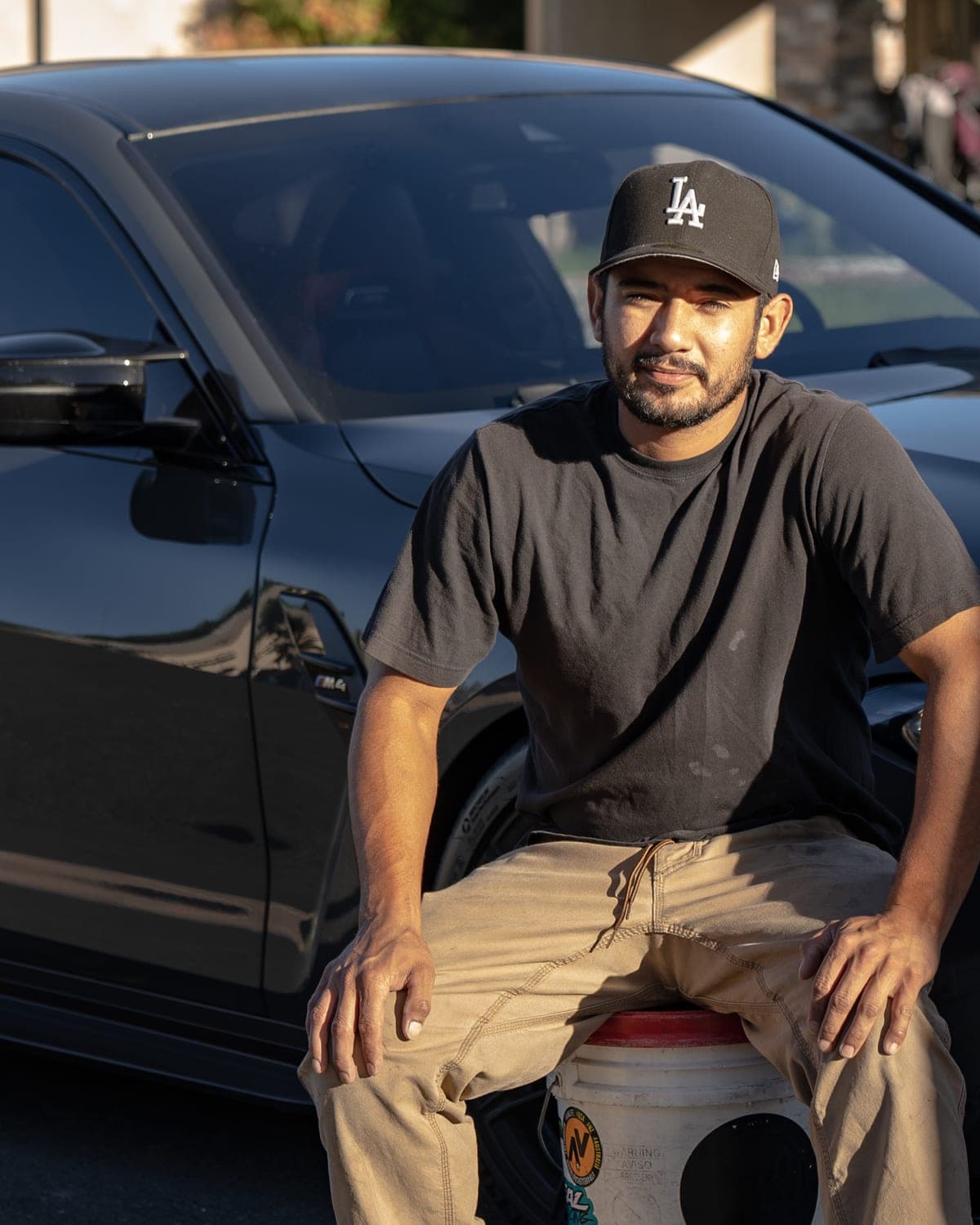 Nick sitting on a detailing bucket beside a black BMW M4 in a Temecula driveway